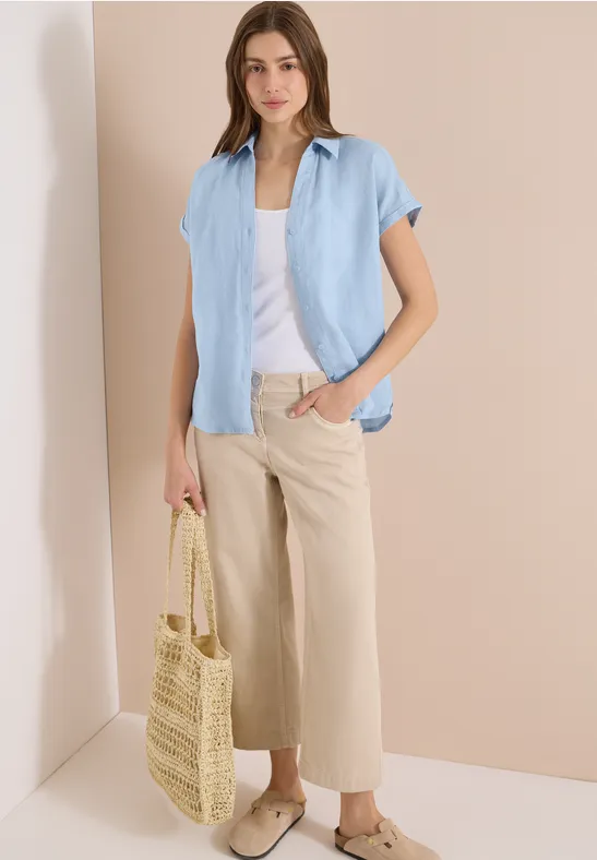 Frau in hellblauer Bluse, weißem Top, beigen Culottes und Strohtasche.