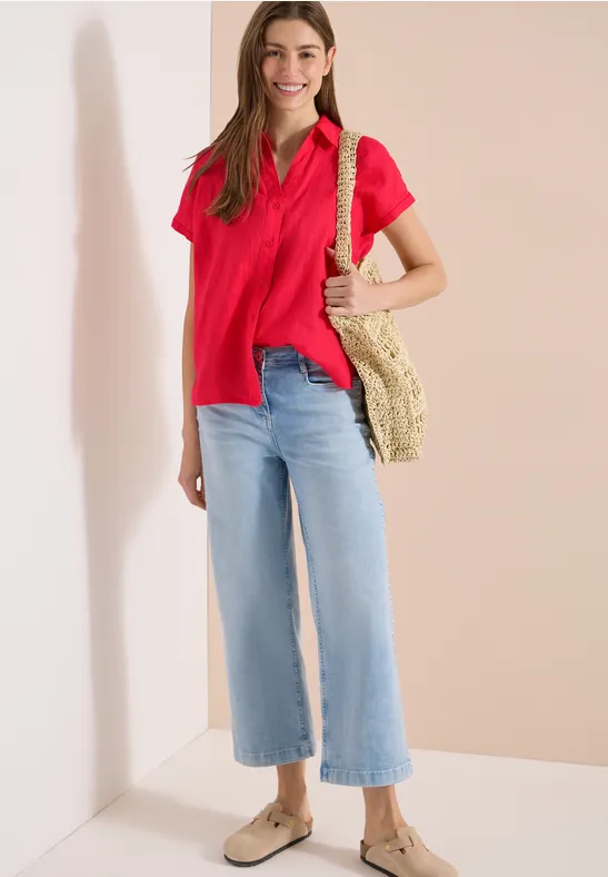 Frau in rotem Poloshirt, hellblauen weiten Chino-Jeans, Basttasche und beigen Espadrilles vor hellem Hintergrund.