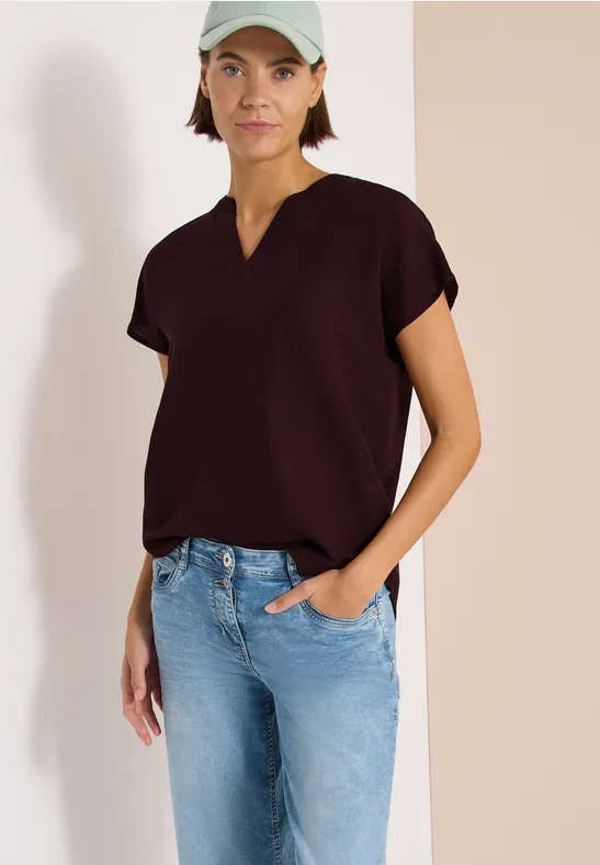 Frau in weinrotem V-Ausschnitt-Shirt und hellblauen Jeans, Hand in Tasche vor hellem Hintergrund.