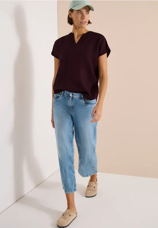 Frau trägt dunkelviolettes V-Neck-T-Shirt, weite 3/4-Jeans und beige Sneakers vor neutralem Hintergrund.