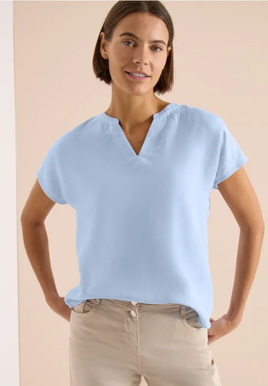 Frau trägt hellblaues kurzärmeliges V-Ausschnitt-Oberteil und beigefarbene Hose, Hände in den Taschen.