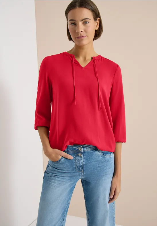 Frau trägt rote Bluse mit Kapuze und hellblaue Jeans, steht vor neutralem Hintergrund.