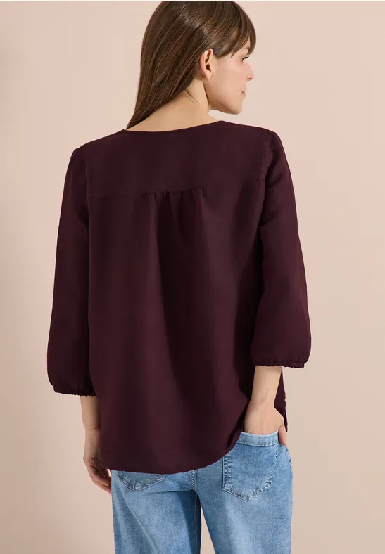 Rückenansicht einer Frau in weinrotem Oberteil mit 3/4-Ärmeln und Jeans vor beigem Hintergrund.