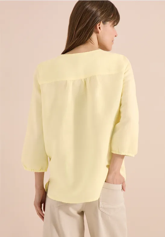 Frau steht mit dem Rücken zur Kamera, trägt eine hellgelbe Bluse mit 3/4-Ärmeln und helle Hosen vor neutralem Hintergrund.