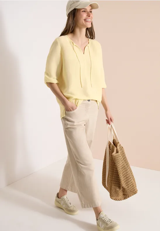 Frau in gelber Bluse, hellen Palazzo-Hosen, Sneakers und Strohtasche vor neutralem Hintergrund.