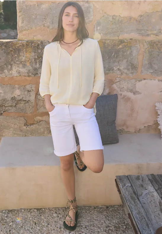 Jemand in hellem Longsleeve und weißen Shorts lehnt entspannt an einer Steinmauer mit einem Bein auf der Bank.