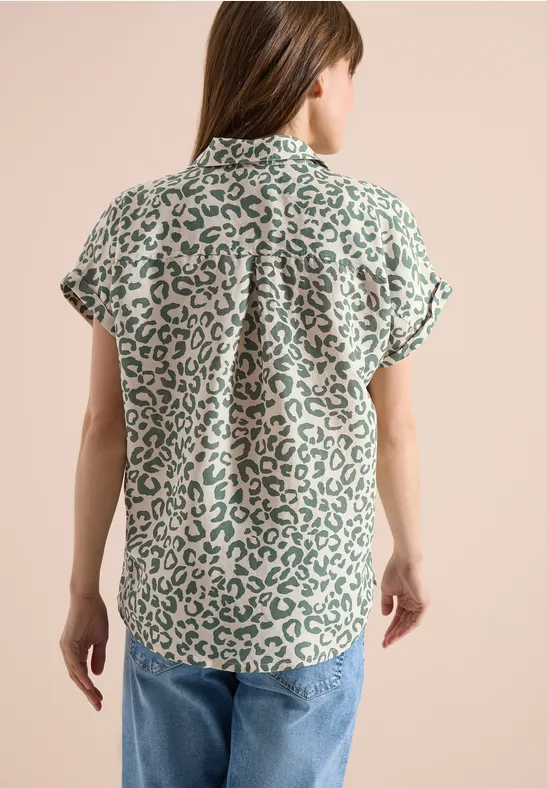 Frau von hinten in kurzärmliger, grün-weißer Leo-Bluse und Jeans vor neutralem Hintergrund.