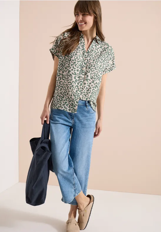 Dame trägt grün gemustertes Kurzarmhemd, blaue Jeans, beige Loafers und hält eine dunkelblaue Tasche.