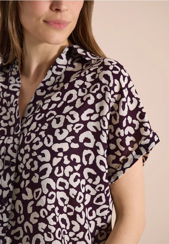 Frau in kurzärmligem, lila-weißem Hemd mit Leopardenmuster vor neutralem Hintergrund.