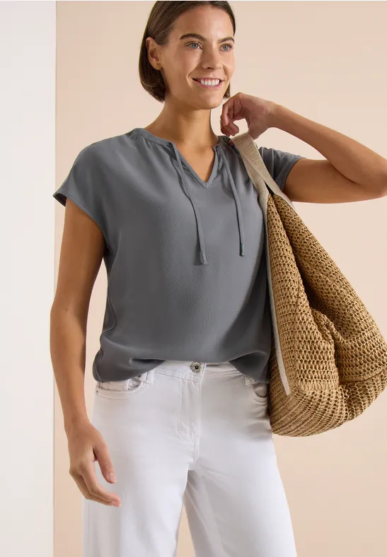 Frau in grauer Bluse, weißen Jeans, mit großer Stroh-Tasche über der Schulter vor beigem Hintergrund.