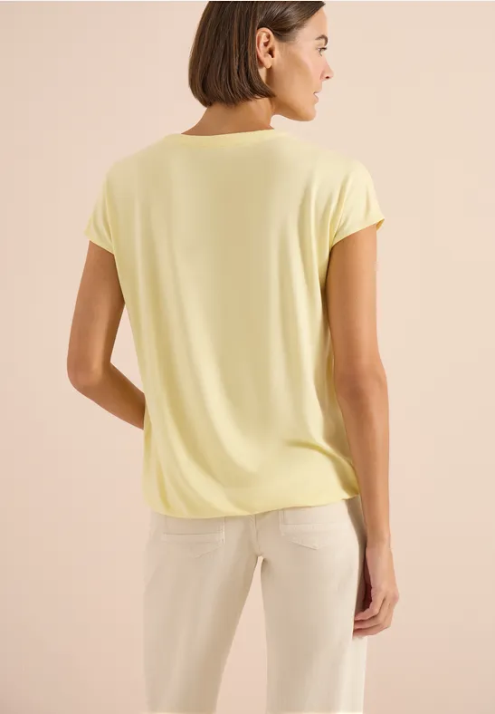 Persoon van achteren met lichtgeel shirt en lichte broek voor een beige achtergrond.