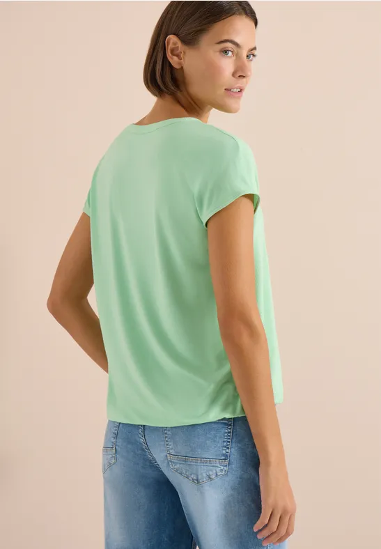 Vrouw in groen T-shirt en blauwe jeans, rugzicht, kijkt over haar schouder.