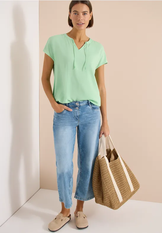Vrouw in groen t-shirt en blauwe jeans met grote strooien tas, staand tegen een lichte muur.