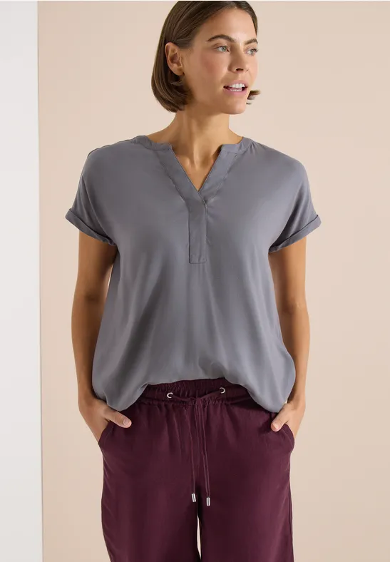 Frau trägt graublaue Bluse mit V-Ausschnitt und weinrote Hose mit Kordel, steht mit Händen in den Taschen.