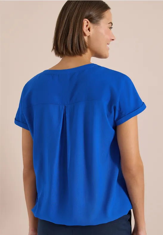 Rückenansicht einer Frau mit kurzem braunen Haar in leuchtend blauer Bluse vor hellem Hintergrund.