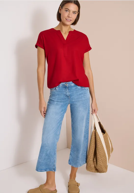 Frau in rotem Kurzarmshirt, hellblauen Jeans-Culottes und Korbtasche vor neutralem Hintergrund.