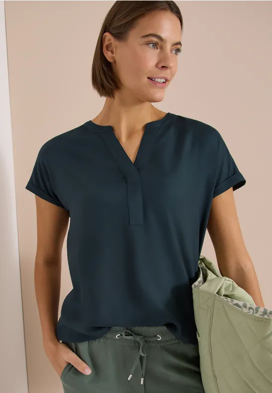 Frau trägt dunkel-türkises Kurzarmshirt mit V-Ausschnitt, olivgrüne Hose mit Kordelzug und passende Tasche.