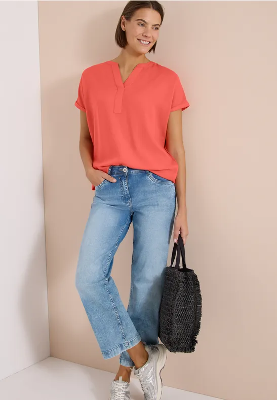 Model trägt korallrotes Shirt, blaue Jeans, weiße Sneaker und hält eine schwarze Flecht-Tasche.