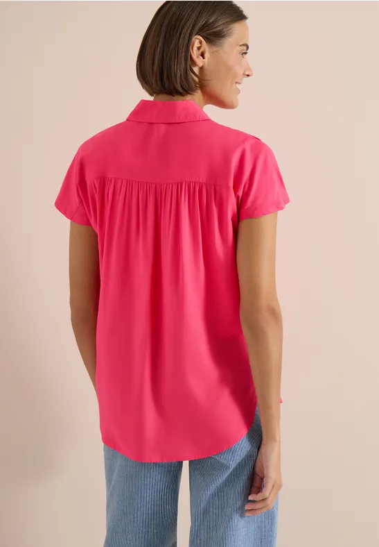 Vrouw van achteren in een felroze blouse met korte mouwen en kraag, gecombineerd met een spijkerbroek.