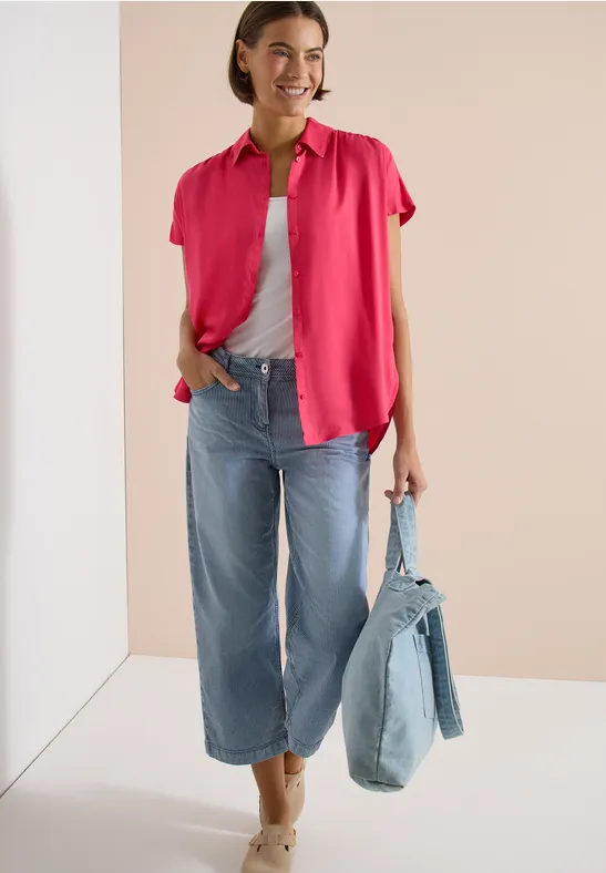 Vrouw met roze blouse over grijs topje, wijde jeans en een denim tas in haar hand.