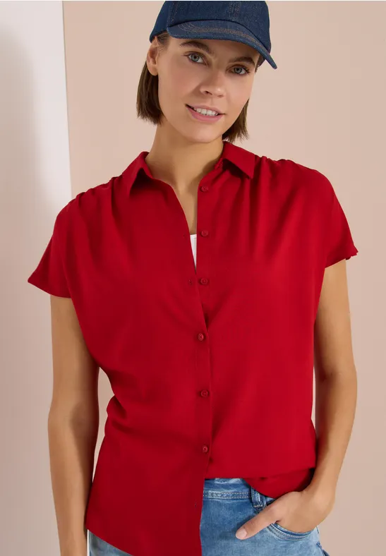 Personne porte une chemise rouge à manches courtes et un jean bleu, une main dans la poche, casquette noire.