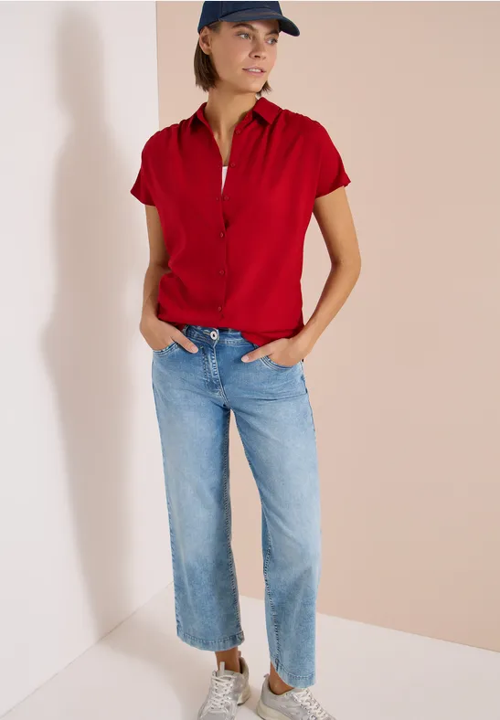 Femme en polo rouge et jean bleu, mains dans les poches, posant détendue dans un coin sur fond clair.