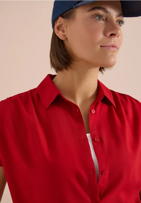 Buste d’une personne portant une chemise rouge, un top blanc et une casquette bleue sur fond neutre.