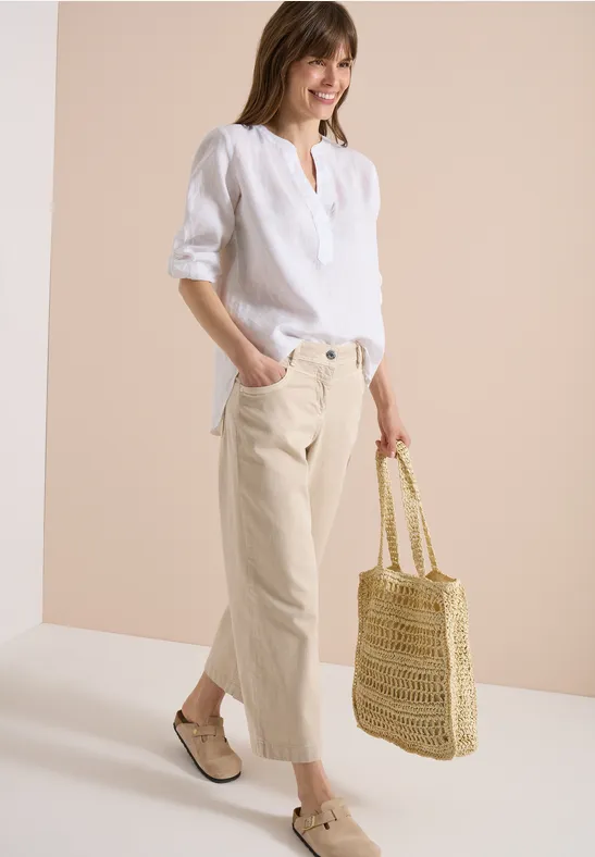 Frau in weißer Leinenbluse, beigen Cropped-Hosen, geflochtenen Espadrilles und Strohtasche vor hellem Hintergrund.