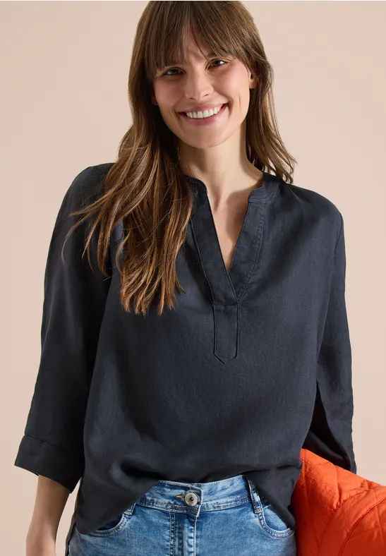 Eine Frau trägt eine dunkelblaue Bluse und blaue Jeans vor einem beigen Hintergrund