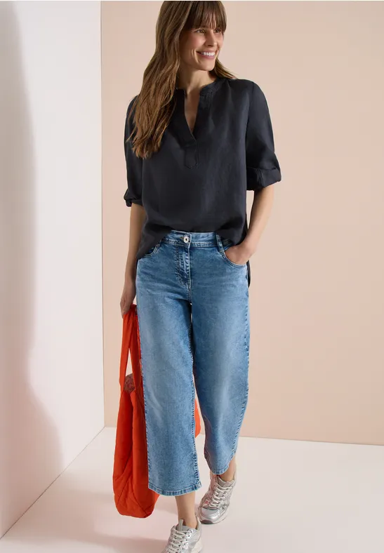Frau trägt schwarze Bluse, weite Jeans, weiße Sneaker und eine orangefarbene Tasche vor neutralem Hintergrund.