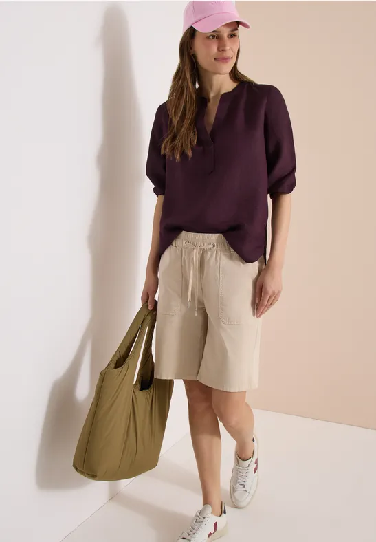 Frau trägt lila Bluse, beige Bermudashorts, weiße Sneaker und große grüne Tasche vor neutralem Hintergrund.