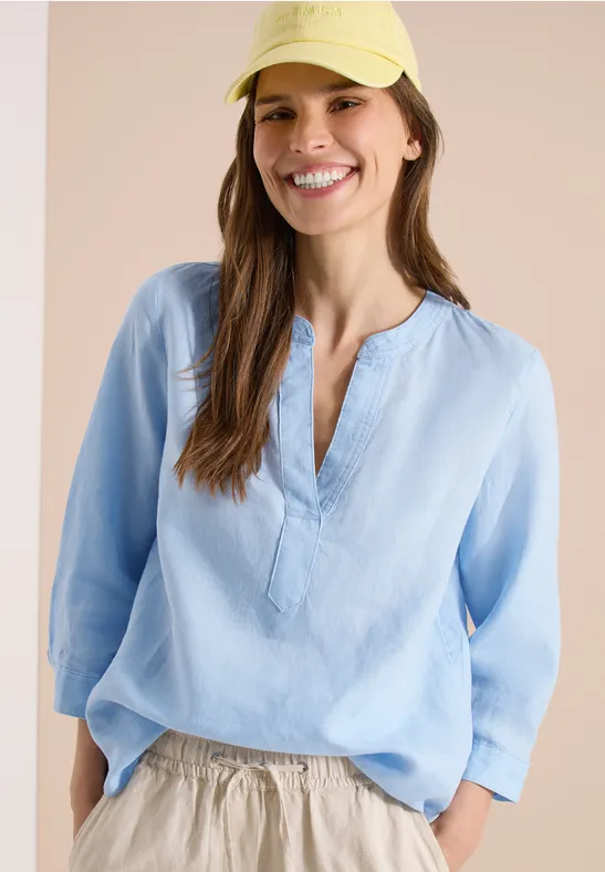 Eine Frau in hellblauer Tunika-Bluse und hellen Hosen, lässiges Outfit mit V-Ausschnitt, Hüfttasche und gelbem Hut.