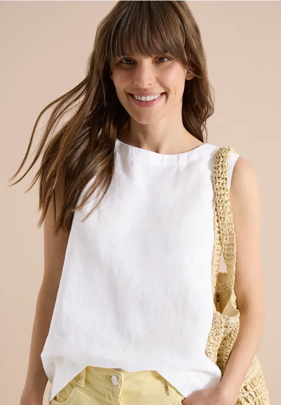 Frau trägt ärmelloses weißes Leinenoberteil und gehäkelte Schultertasche vor neutralem Hintergrund.