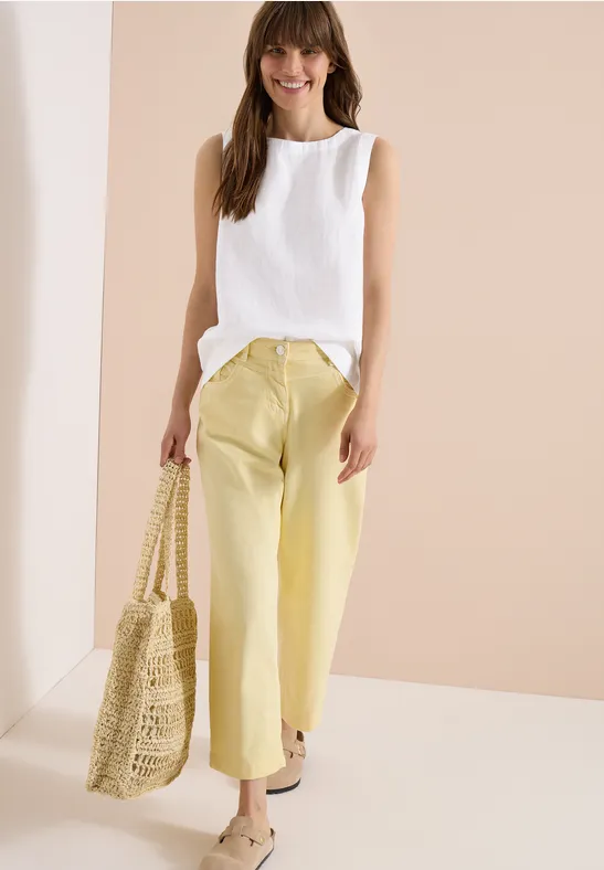 Frau in weißem ärmellosem Top, gelben Hosen und geflochtener Tasche, vor hellem Hintergrund