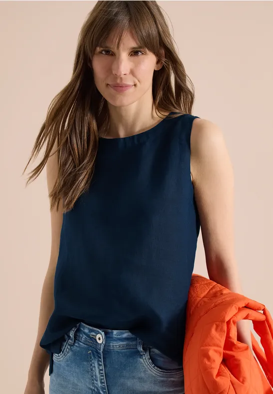 Femme en top sans manches bleu marine et jean, tenant une veste orange à la main.
