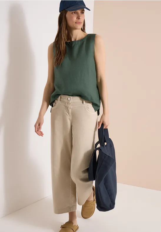 Frau mit grünem Top und beigen Weitbein-Hosen, trägt Mütze und dunkelblaue Tasche.