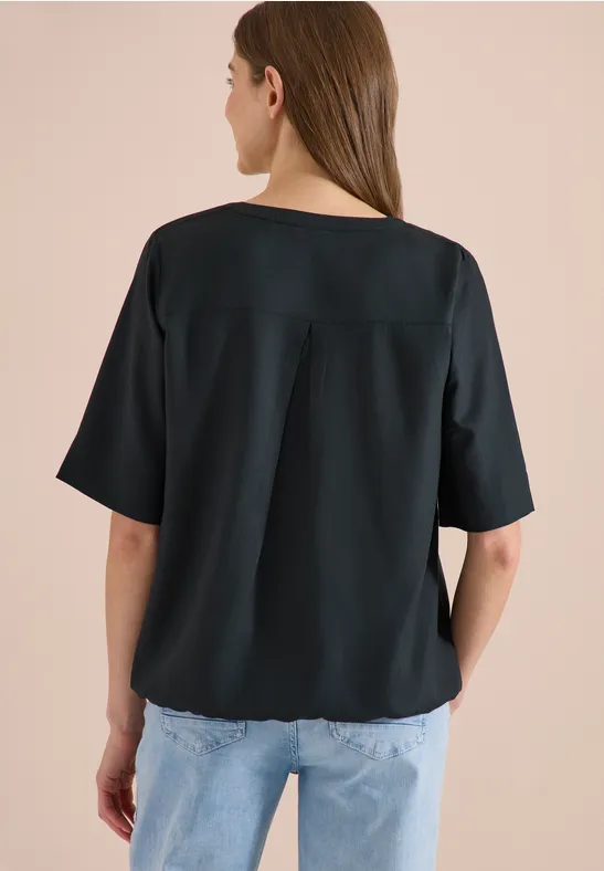 Rückansicht einer Frau in lockerem schwarzen Kurzarmshirt und blauen Jeans vor beigem Hintergrund.