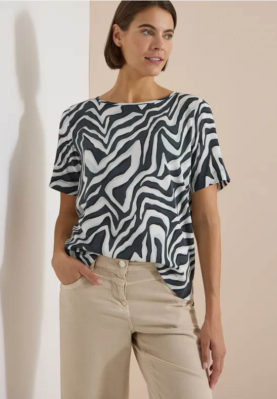 Frau trägt ein kurzärmeliges Zebra-Muster-Oberteil und beige Hose vor neutralem Hintergrund.