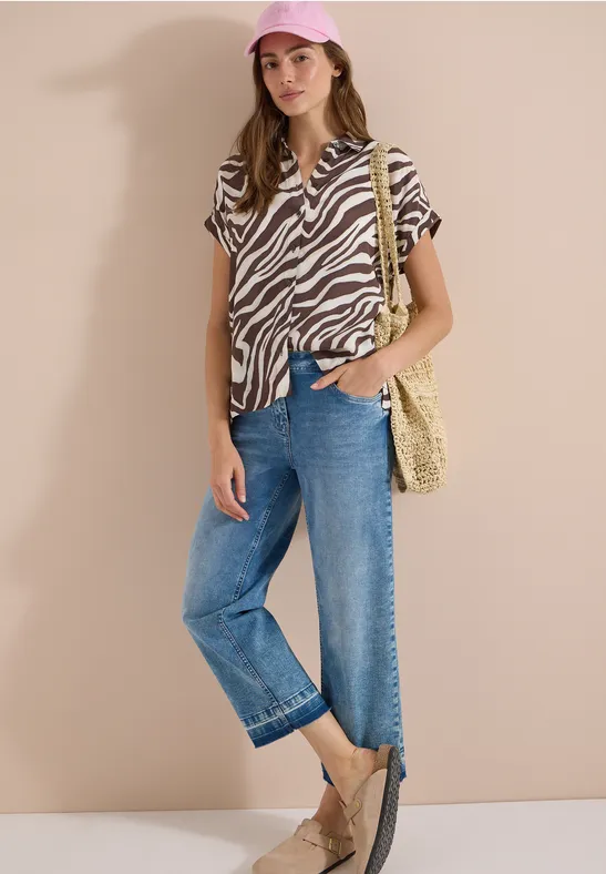 Vrouw in zebra-print blouse, wijde jeans, strooien tas, espadrilles en pet voor een beige muur.
