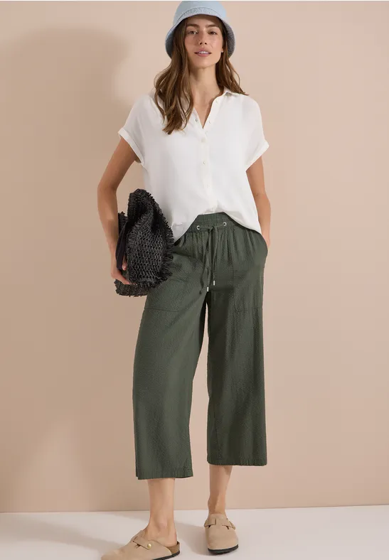 Femme en chemisier blanc, pantalon culotte olive, mocassins beiges et sac tressé devant un mur beige.