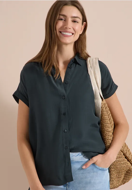 Femme en chemise courte vert foncé, manches retroussées, sac en osier au bras, main dans la poche, fond clair.