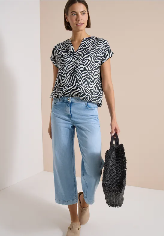 Frau mit zebragemustertem Oberteil, weiten Jeans, Basttasche und Espadrilles.