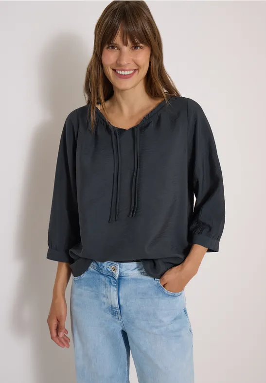 Lächelnde Frau trägt dunkle Bluse mit Kordeln und helle Jeans, steht entspannt vor heller Wand.