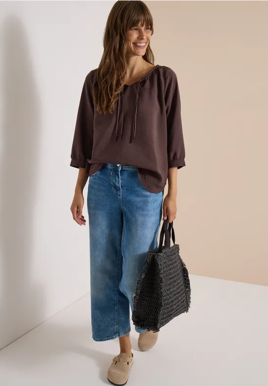 Frau in brauner Bluse, weiten Jeans und Sneakers trägt schwarze geflochtene Tasche und lächelt.
