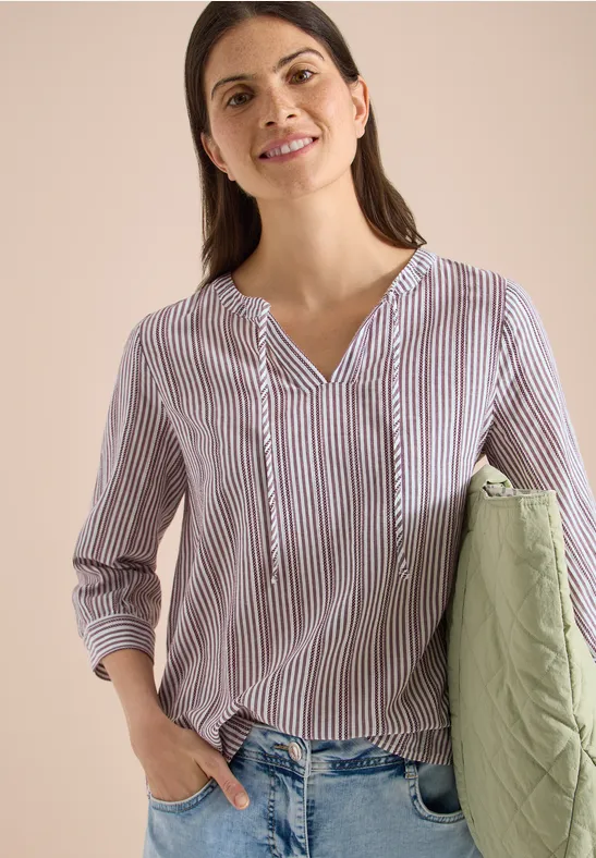 Frau trägt gestreifte Bluse mit V-Ausschnitt und 3/4-Ärmeln, Jeans, hält grüne gesteppte Tasche.