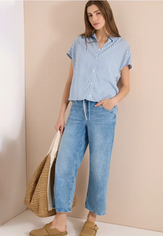 Frau in blau-weiß gestreiftem Kurzarmhemd, weiten Jeans und Strohtasche vor beigem Hintergrund.