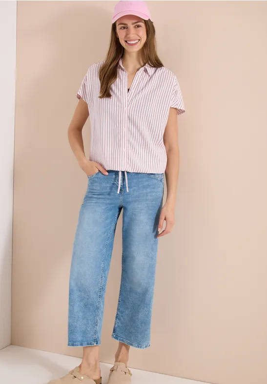 Frau in gestreiftem Hemd, weiten Jeans und Sneakers vor beigem Hintergrund.