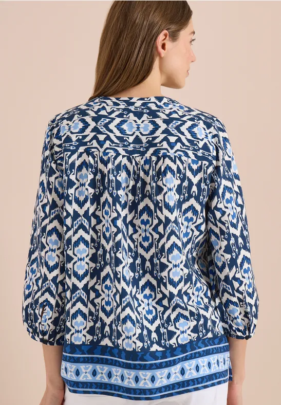 Frau von hinten in blau-weiß gemustertem, locker sitzendem Oberteil mit 3/4-Ärmeln vor neutralem Hintergrund.