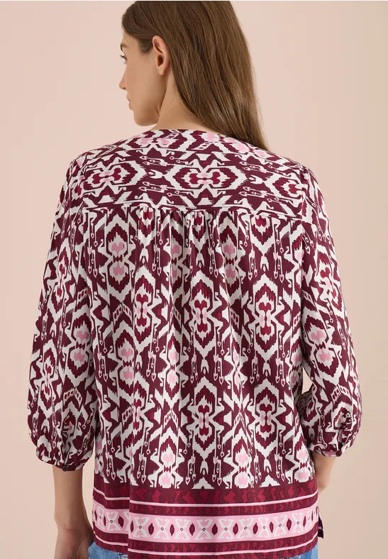 Rückansicht einer Frau in locker geschnittener, rot-weiß gemusterter Bluse mit 3/4-Ärmeln vor beigem Hintergrund.