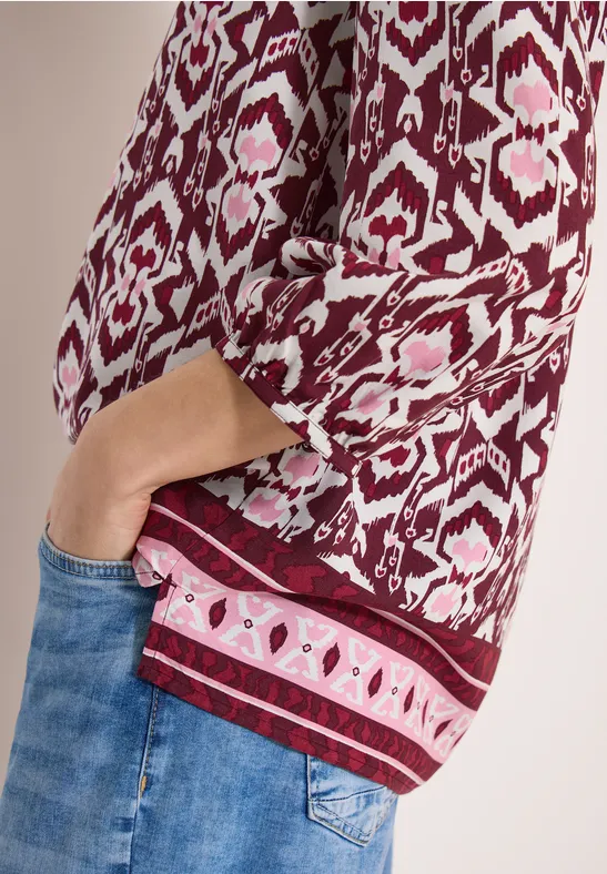 Rote Bluse mit Ethno-Muster, 3/4-Ärmel, Hand in Jeans-Tasche.